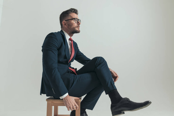 vista lateral de empresário sexy com pernas cruzadas olhando para cima e pensando enquanto sentado em fundo cinza no estúdio - Foto, Imagem