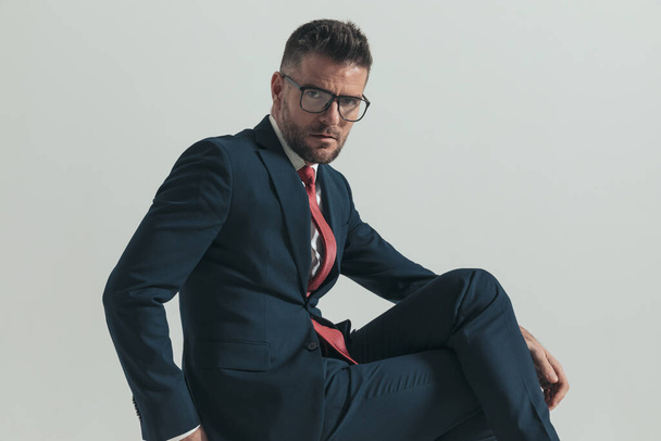 Seitenansicht eines eleganten Typen in den Vierzigern mit Brille, die die Beine kreuzt, während er auf einem Holzstuhl vor grauem Hintergrund im Studio sitzt - Foto, Bild