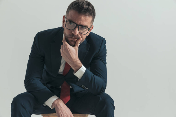pensive young businessman with beard looking away, thinking, holding elbows on knees and sitting on wooden chair in studio - Photo, Image