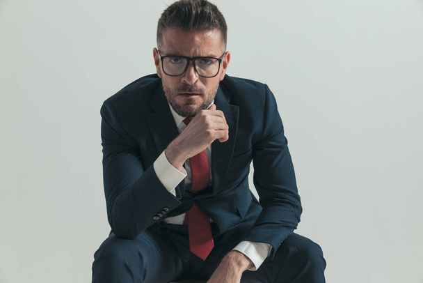 portrait d'homme d'affaires sexy d'âge moyen avec des lunettes tenant les coudes sur les genoux et posant devant fond gris en studio - Photo, image