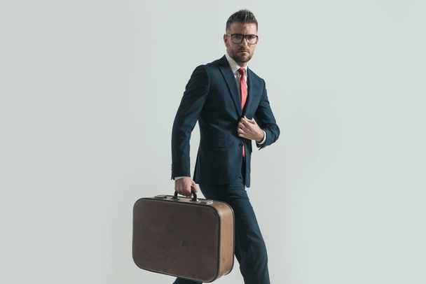handsome bearded man in his forties holding luggage, fixing suit and leaving town while walking on grey background in studio - Fotoğraf, Görsel