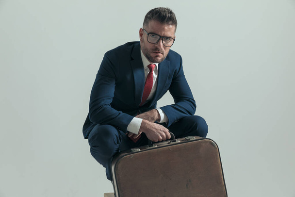 Porträt eines bärtigen Geschäftsmannes mit Gepäck, das Ellbogen auf Knien hält und vor grauem Hintergrund im Studio auf seinen Flug wartet - Foto, Bild