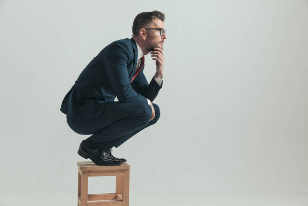 side view of pensive man looking to side and dreaming about perfect life while crouching on wooden chair in studio on grey background - Photo, Image