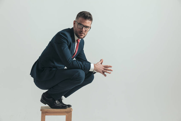 side view of attractive man in his forties crouching on wooden chair and touching palms on grey background in studio - Foto, immagini