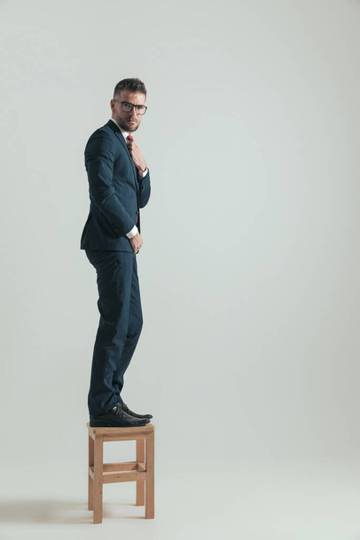 elegante bebaarde zakenman staand op houten stoel en bevestigingsstropdas terwijl poseren in een zijaanzicht pose op grijze achtergrond - Foto, afbeelding