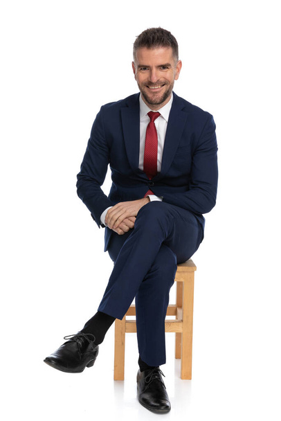 Eleganter Mann in seinen Vierzigern mit Bart, der die Beine kreuzt, während er im Studio auf einem Holzstuhl vor weißem Hintergrund sitzt - Foto, Bild