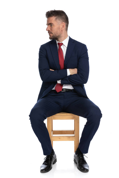 uspet businessman in suit with red tie on wooden chair crossing arms while looking to side in front of white background in studio - Photo, Image