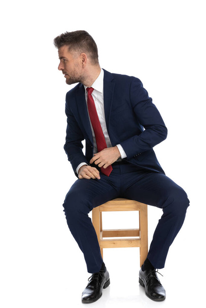 κομψός επιχειρηματίας με κοστούμι ψάχνει για πλευρά με περίεργο τρόπο, ενώ κάθεται μπροστά από το λευκό φόντο στο στούντιο - Φωτογραφία, εικόνα