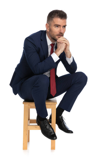 thoughtful man in his forties wearing elegant navy blue suit, looking to side, holding hands together and thinking of ways to solve difficult problems on white background - Foto, afbeelding
