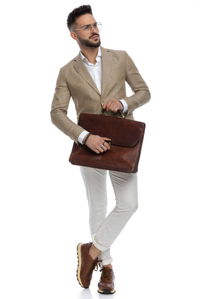 sexy hombre de negocios sosteniendo un maletín y cruzando los pies y mirando hacia otro lado en una pose de moda - Foto, imagen