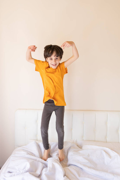 An energetic child is jumping on the bed, a boy in pajamas is fooling around in his room, the child is enjoying the weekend - 写真・画像