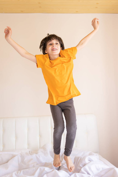 An energetic child is jumping on the bed, a boy in pajamas is fooling around in his room, the child is enjoying the weekend - Foto, Imagen