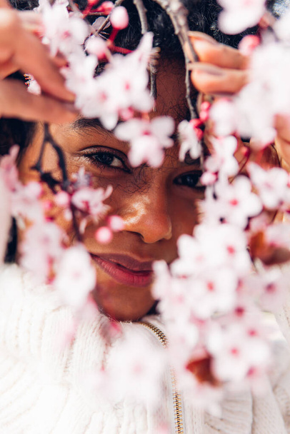 Retrato vertical de una mujer rodeada de flores rosas en primavera. Joven mujer adulta de etnia afroamericana con cabello oscuro y afro, mirando a la cámara en la naturaleza, concepto de pureza y feminidad. - Foto, imagen