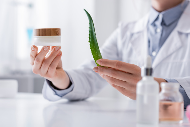 cropped view of laboratory assistant holding aloe and container with cream in laboratory - Foto, Bild