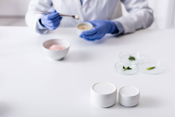 recipientes con crema cerca de asistente de laboratorio borroso en guantes de látex  - Foto, Imagen