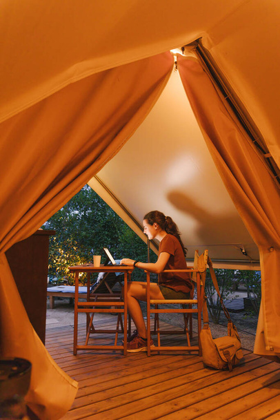 Fénylő sátor egy nővel, aki laptoppal dolgozik alkonyatkor. Luxus kemping sátor szabadtéri nyári életmód koncepció - Fotó, kép