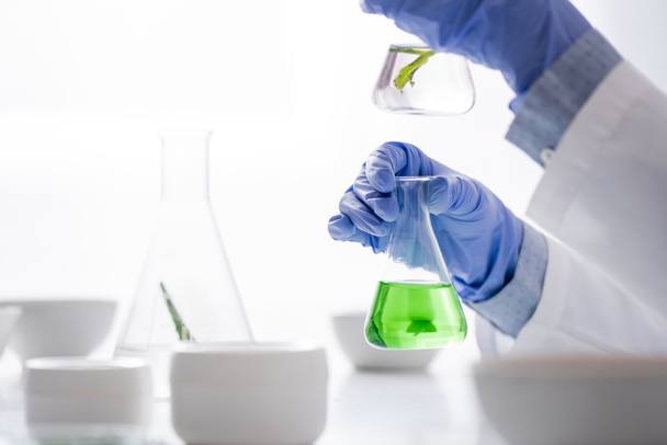 Ausgeschnittene Ansicht einer Laborantin in Latexhandschuhen, die Flaschen mit grüner Flüssigkeit in der Hand hält  - Foto, Bild