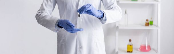 vista recortada del asistente de laboratorio en guantes de látex sosteniendo botella con suero en laboratorio, pancarta - Foto, imagen