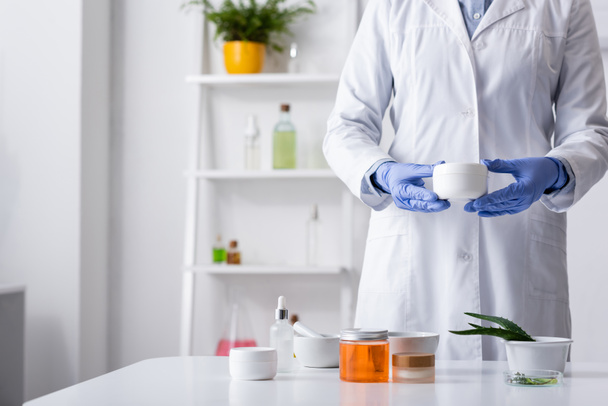 обрезанный вид лаборанта в латексных перчатках, держащего контейнер со сливками рядом с косметическими ингредиентами  - Фото, изображение