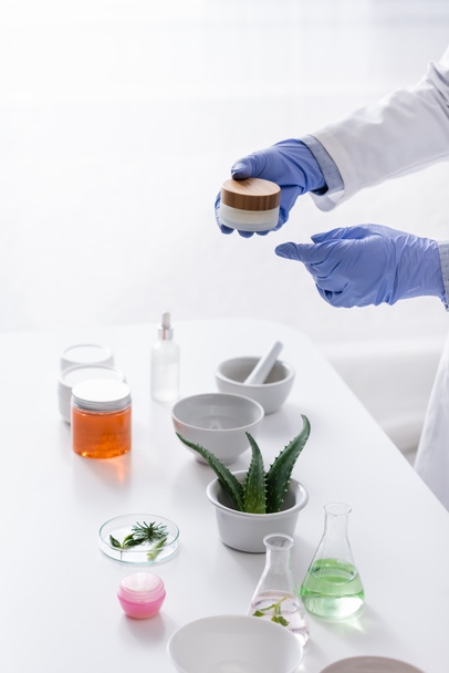partial view of laboratory assistant in latex gloves holding container with cream near aloe and cosmetic ingredients in lab - Photo, Image