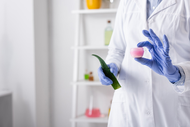 cropped view of laboratory assistant in latex gloves holding aloe leaf and container with cream - Fotoğraf, Görsel