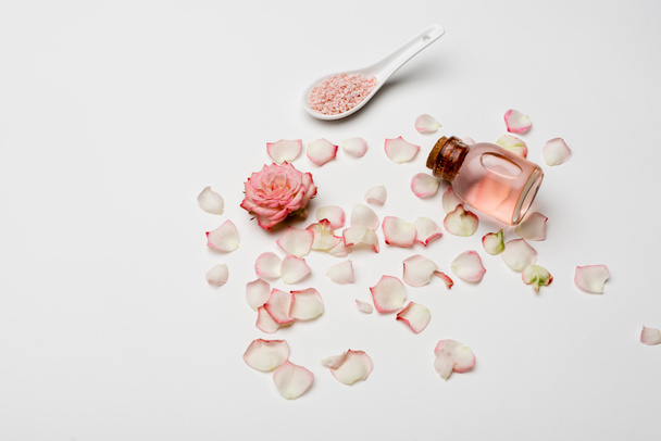 vysoký úhel pohledu růže v blízkosti okvětních lístků, růžová mořská sůl a láhev s růžovou vodou na bílém - Fotografie, Obrázek