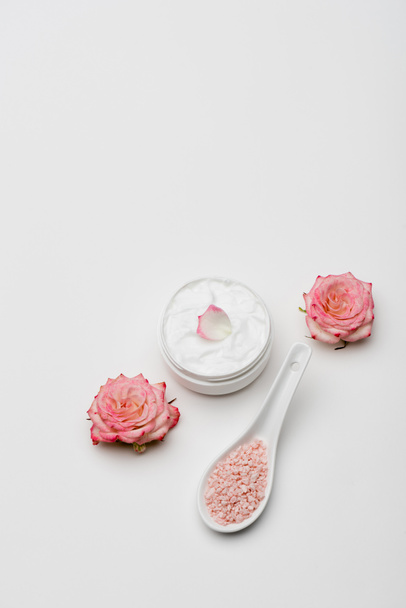 bovenaanzicht van bloemen in de buurt van roze zeezout en container met crème op wit - Foto, afbeelding