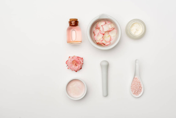 top view of flower near pestle and mortar with petals, sea salt, cream and bottle with rose water on white - Foto, immagini