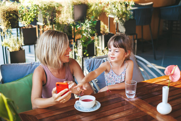 Blonde woman and her little daughter using smartphone for video chat in the cafe. Lifestyle, technology and communication concept. - Photo, Image