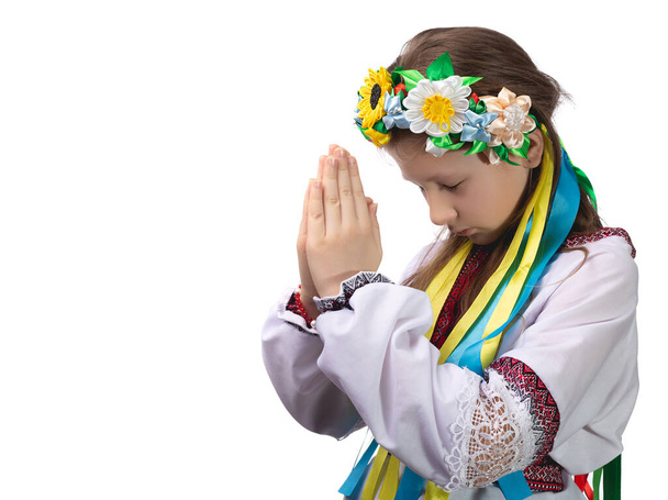 涙の目をしたウクライナの民族衣装を着た少女が彼女の前で手を折り、ウクライナでの戦争の終わりを祈ります。白い背景のスタジオ写真. - 写真・画像