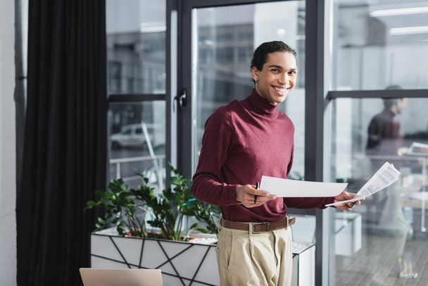 Χαμογελώντας αφροαμερικανός επιχειρηματίας κρατώντας χαρτιά κοντά σε φορητό υπολογιστή στο γραφείο  - Φωτογραφία, εικόνα
