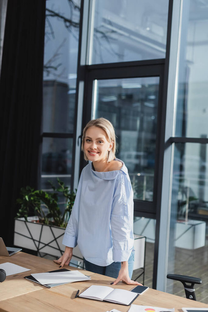 Positive businesswoman looking at camera near documents and devices in office  - Valokuva, kuva
