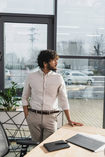 Side view of young indian businessman standing near working table in office  - Foto, immagini