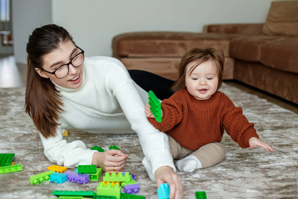 ベビーシッターと幼児の女の子は自宅でおもちゃの色のブロックで遊んでいます。茶色のセーターの少女。眼鏡をかけた女性。幸せな家族は一緒に時間を過ごす. - 写真・画像