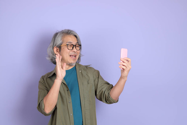 The senior Asian man with casual clothes standing on the purple background. - Photo, image