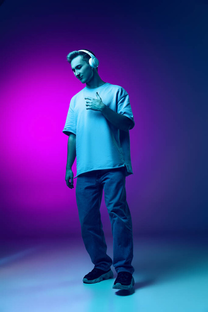Full-length portrait of young emotive man listening to music in headphones isolated over gradient pink and purple background in neon - Valokuva, kuva