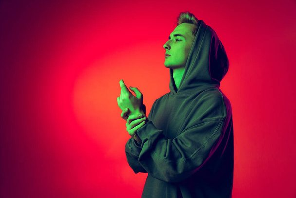 Portrait of stylish young man in big hoodie posing isolated over red studio background in green neon light - Foto, Imagem