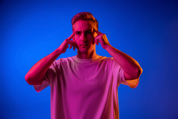 Portrait of young emotive man touching temples with thoughtful expression isolated over blue studio background in pink neon light - Foto, Bild
