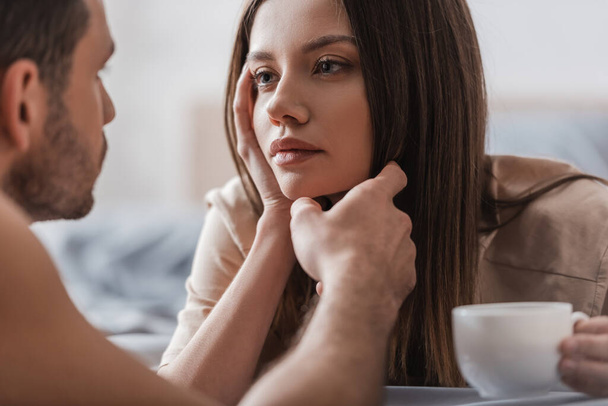 Blurred man touching face of brunette girlfriend with cup of coffee in bedroom  - Φωτογραφία, εικόνα