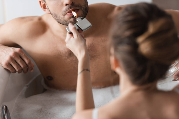 Молодая женщина держит зажигалку рядом с мускулистым парнем с сигаретой в ванной  - Фото, изображение