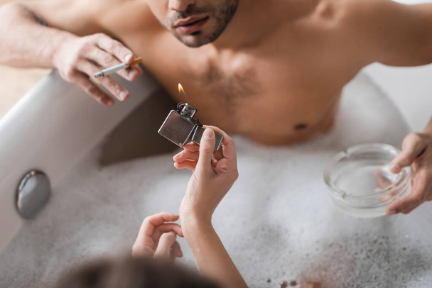 Overhead view of woman holding lighter near blurred boyfriend with cigarette and ashtray in bathtub  - Foto, imagen
