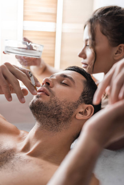 Sexy man holding cigarette near blurred girlfriend with ashtray in bathtub  - 写真・画像