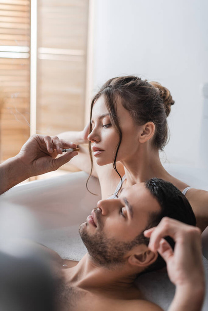 Bearded man holding cigarette near girlfriend in bathtub with foam  - 写真・画像