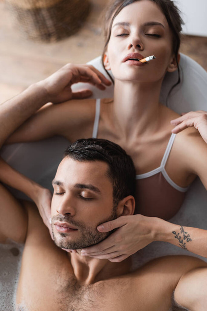 Top view of brunette woman with closed eyes smoking and touching bearded boyfriend in bath  - Фото, изображение