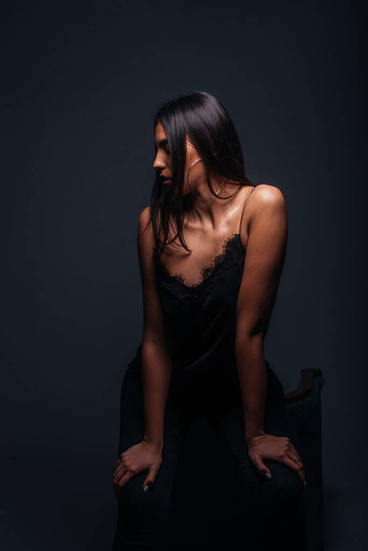 Nádherný ženský model se sexy černou tílko top a černé kalhoty pózující před židlí uvnitř fotostudia - Fotografie, Obrázek