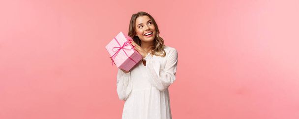 Fiestas, celebración y concepto de mujer. Retrato de la joven rubia atractiva curiosa celebrando cumpleaños, sacudiendo caja de regalo rosa para averiguar lo que está dentro, sonriendo cámara mirada intrigada
 - Foto, imagen
