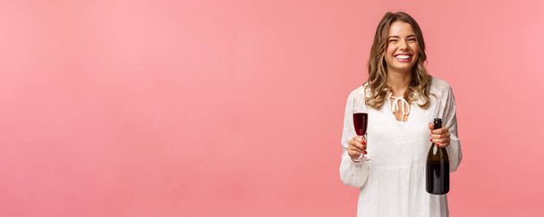Vacaciones, primavera y concepto de fiesta. Retrato de mujer rubia europea feliz y despreocupada celebrando en vestido blanco, sosteniendo botella de champán o vino, bebiendo de copa y riendo
 - Foto, Imagen
