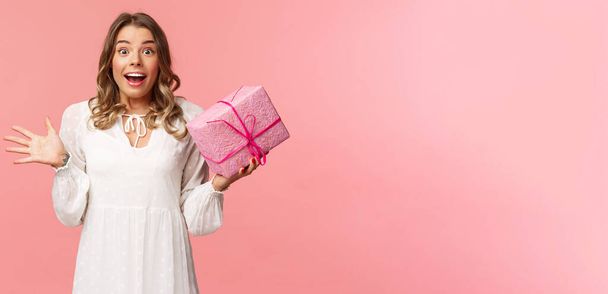 Holidays, celebration and women concept. Portrait of surprised upbeat charismatic blond girl in white dress, spread hands sideways and gasping amazed, receive surprise gift, hold pink box - Foto, Bild
