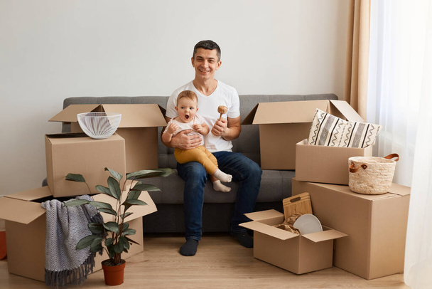 Portrait of young adult brunette man wearing white t shirt sitting on sofa with baby daughter during moving to a new flat, posing surrounded with cardboard box. - Zdjęcie, obraz