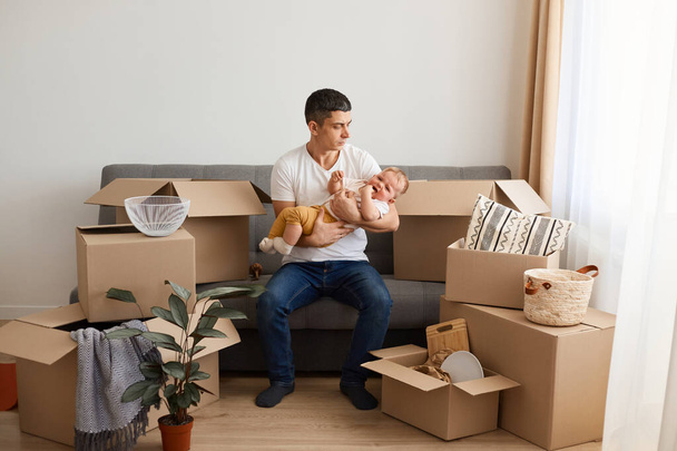 Portrait of brunette man wearing white t shirt sitting on sofa with baby daughter during moving to a new house, posing with infant, trying to calm down his child. - Valokuva, kuva